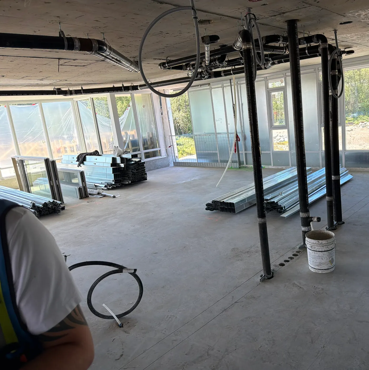 Work area on a concrete floor: hanging pipe and conduit, stacked steel studs, window frames leaning against a wall, a coiled hose on the floor and a guardrail at the window opening.
